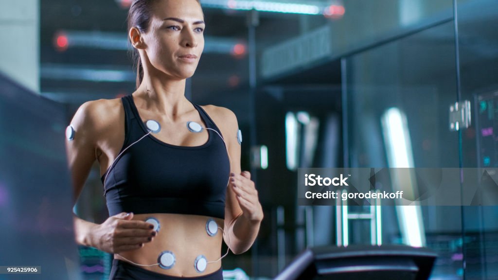 Beautiful Woman Athlete Runs on a Treadmill with Electrodes Attached to Her Body, Female Physician Uses Tablet Computer and Controls EKG Data Showing on Laboratory Monitors. Adult Stock Photo