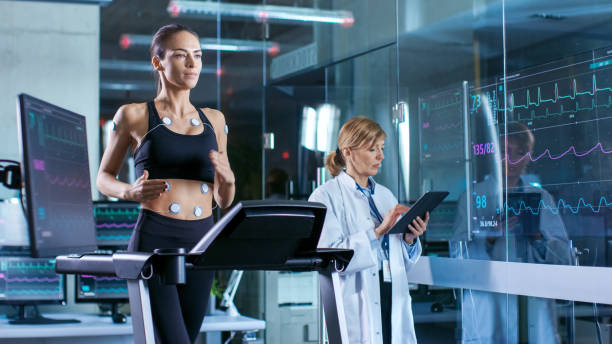 hermosa mujer atleta corre en una caminadora con electrodos conectados a su cuerpo, médico femenino utiliza el tablet pc y controla datos de electrocardiograma que muestra en los monitores de laboratorio. - human heart surveillance computer monitor pulse trace fotografías e imágenes de stock