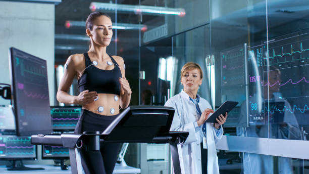 beautiful woman athlete runs on a treadmill with electrodes attached to her body, female physician uses tablet computer and controls ekg data showing on laboratory monitors. - smiling research science and technology clothing imagens e fotografias de stock