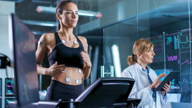 Beautiful Woman Athlete Runs on a Treadmill with Electrodes Attached to Her Body, Female Physician Uses Tablet Computer and Controls EKG Data Showing on Laboratory Monitors. Beautiful Woman Athlete Runs on a Treadmill with Electrodes Attached to Her Body, Female Physician Uses Tablet Computer and Controls EKG Data Showing on Laboratory Monitors. biomechanics stock pictures, royalty-free photos & images