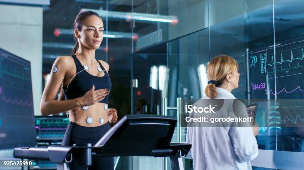 Beautiful Woman Athlete Runs On A Treadmill With Electrodes Attached To Her Body Female Physician Uses Tablet Computer And Controls Ekg Data Showing On Laboratory Monitors Stock Photo - Download Image Now