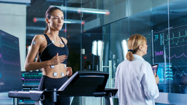 beautiful woman athlete runs on a treadmill with electrodes attached to her body, female physician uses tablet computer and controls ekg data showing on laboratory monitors. - smiling research science and technology clothing imagens e fotografias de stock