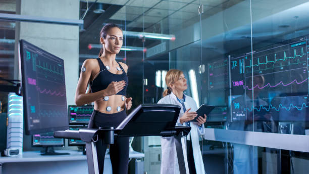 hermosa mujer atleta corre en una caminadora con electrodos conectados a su cuerpo, médico femenino utiliza el tablet pc y controla datos de electrocardiograma que muestra en los monitores de laboratorio. - sistema de conducción cardíaco fotografías e imágenes de stock