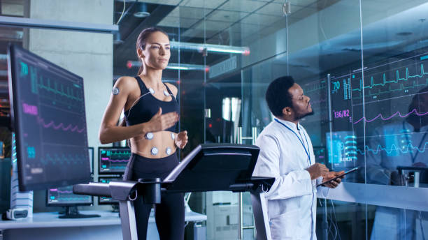 beautiful woman athlete runs on a treadmill with electrodes attached to her body, physician uses tablet computer and controls ekg data showing on laboratory monitors. - smiling research science and technology clothing imagens e fotografias de stock