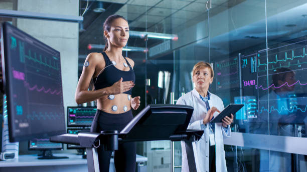 hermosa mujer atleta corre en una caminadora con electrodos conectados a su cuerpo, médico femenino utiliza el tablet pc y controla datos de electrocardiograma que muestra en los monitores de laboratorio. - human heart surveillance computer monitor pulse trace fotografías e imágenes de stock