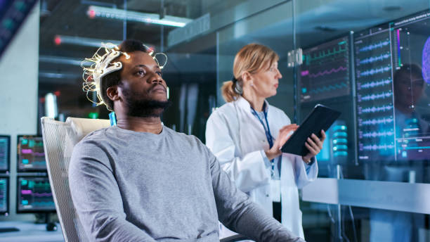 istock In Laboratory Man Wearing Brainwave Scanning Headset Sits in a Chair while Scientist with Tablet Computer Supervises Process. In the Modern Brain Study/ Neurological Research Laboratory. Monitors Show EEG Reading and Graphical Brain Model. 925426094