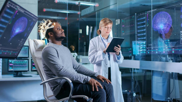 istock Man Wearing Brainwave Scanning Headset Sits in a Chair while Scientist with Tablet Computer Supervises Process. In the Modern Brain Study Laboratory Monitors Show EEG Reading and Brain Model. 925426154
