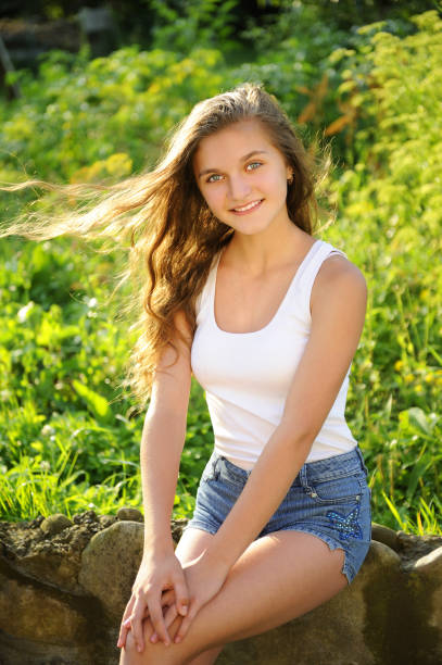 A cheerful girl sits on a nature background. The setting sun shines on her, and a light warm summer wind blows her long hair. She is wearing a white T-shirt and short jeans shorts. She smiles. She likes to be photographed.