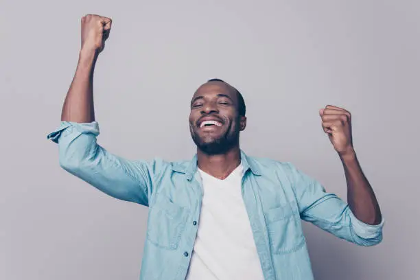 Dreams come true! Portrait of excited cheerful handsome delightful joyous wearing casual denim shirt guy raising his hands up, isolated on grey background