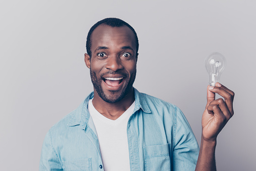 Hooray! Close up portrait of excited cheerful glad delightful handsome shocked amazed astonished man wearing denim jeans shirt holding light bulb isolated on gray background