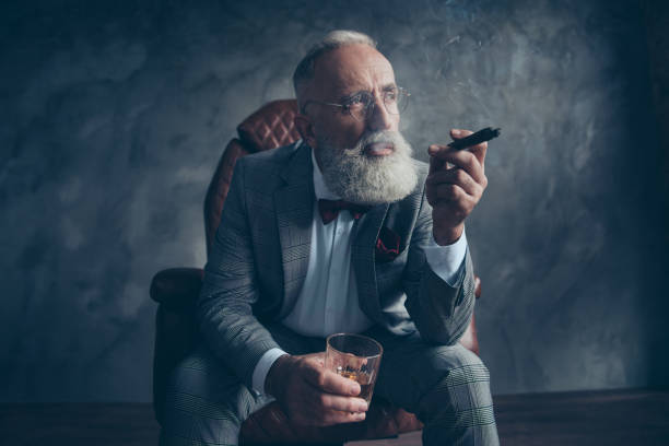 mentalità, meditare uomo ricco in bicchieri, sigaretta fumo, fumi, tenere il vetro con brandy, in smoking con papillon rosso, sedersi in poltrona di pelle su sfondo grigio, guardando di lato, azioni, azioni, soldi - cigar whisky men smoking foto e immagini stock