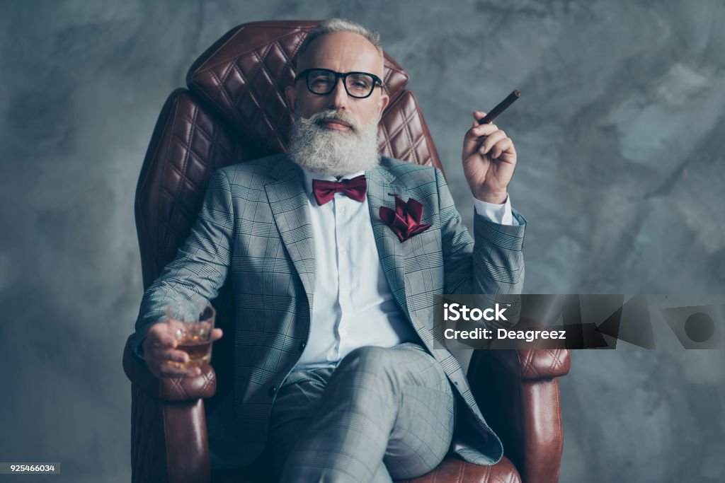 Cool man en vasos, sostener el cigarrillo, vidrio con brandy, en ropa formal, tux con pajarita roja y Plaza de bolsillo, sentarse en la silla de cuero sobre fondo gris, mirando a la cámara, acciones, valores, dinero - Foto de stock de Hombres libre de derechos