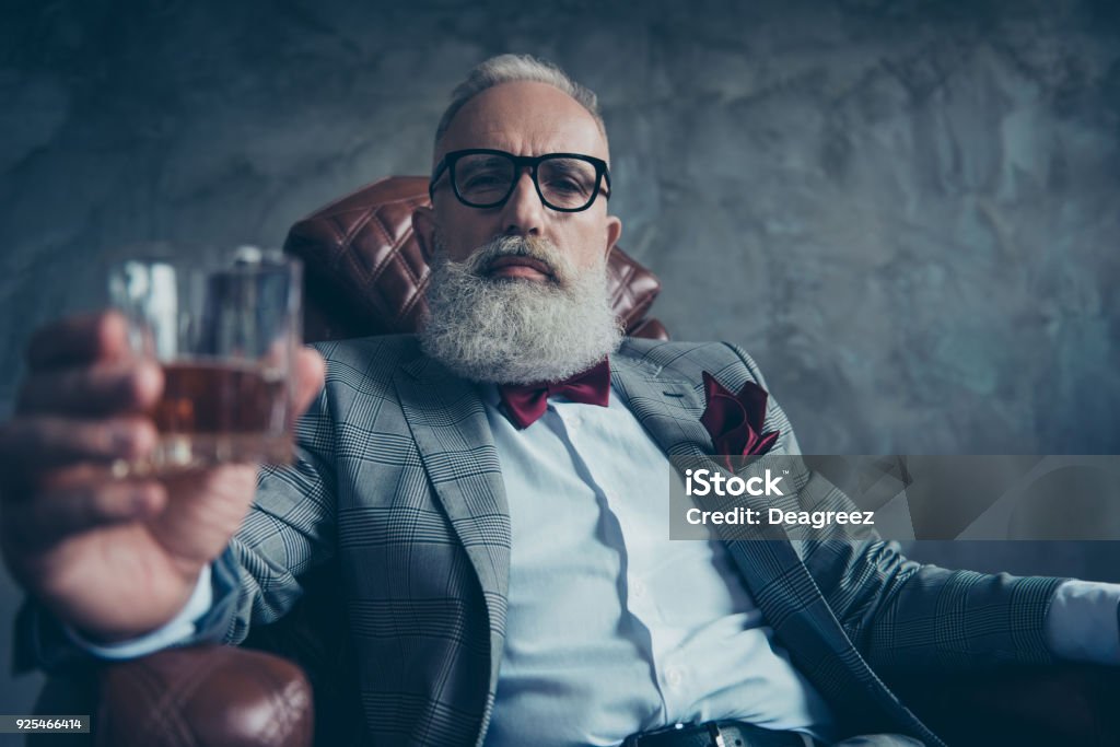 Attraente, vecchio investitore in occhiali, tenere il vetro con brandy, in smoking con papillon rosso e quadrato tascabile, sedersi su sedia di pelle su sfondo grigio, guardando la fotocamera, azioni, azioni, denaro - Foto stock royalty-free di Whisky