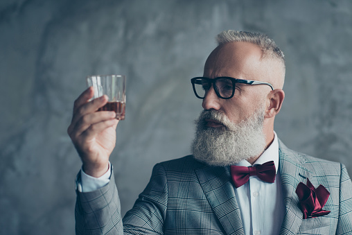 Close up photo of serious pensive confident satisfied bearded rich wealthy entrepreneur wine-maker manufacturer looking at the glass with whisky, isolated on grey background