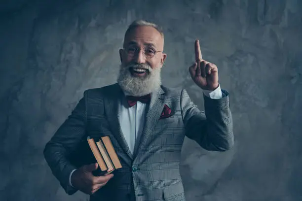 Portrait  of successful, clever attractive, serious millionaire in jacket with bow-tie, in glasses, holding three books in arms, pointing forefinger up, find a great idea over gray background