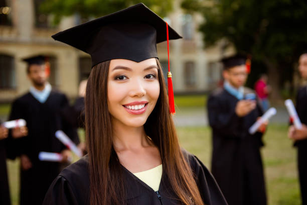 felice ragazza bruna cinese carina sta sorridendo, compagni di classe sfocati con diplomi dietro. lei è in una tavola di mortaio nero, con nappa rossa, in abito, capelli castani, sorriso bianco dento, così eccitato - graduation student women beauty foto e immagini stock