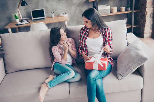 Happy women's day dear mummy! Little lovely beautiful daughter is waiting her mum to open a red giftbox from her, they are sitting on a sofa at home, domestic cozy atmosphere