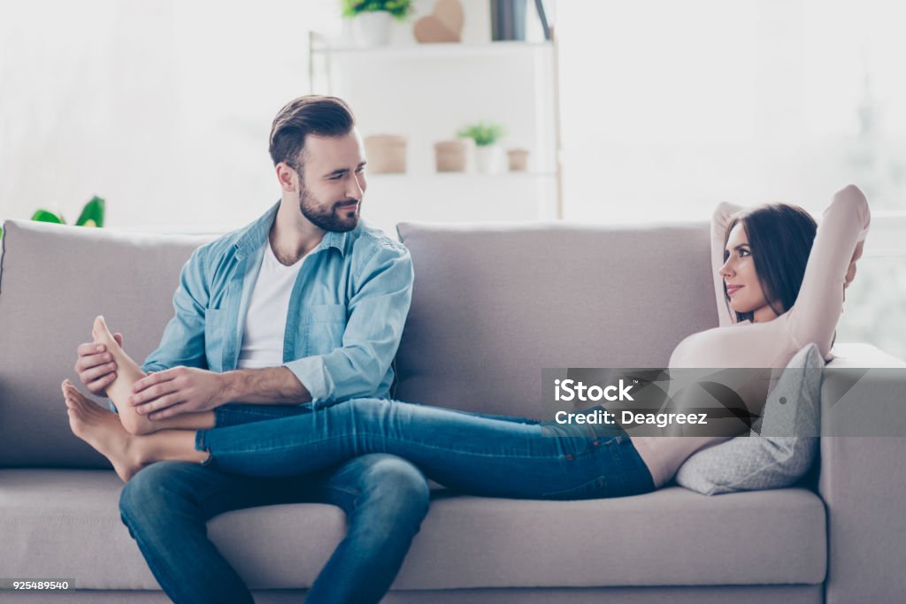 I bliss out and on cloud nine! Attentive caring loving handsome bearded husband is doing feet massage for her attractive tender lying on a sofa wife Massaging Stock Photo