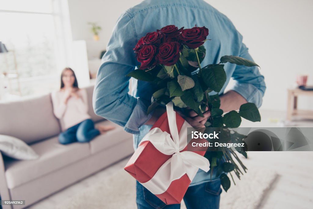 Unexpected moment in routine everyday life! Cropped photo of man's hands hiding holding chic bouquet of red roses and gift with white ribbon behind back, happy woman is on blurred background Valentine's Day - Holiday Stock Photo