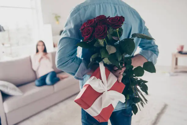 Photo of Unexpected moment in routine everyday life! Cropped photo of man's hands hiding holding chic bouquet of red roses and gift with white ribbon behind back, happy woman is on blurred background