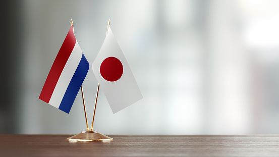 Japanese and Dutch flag pair on desk over defocused background. Horizontal composition with copy space and selective focus.