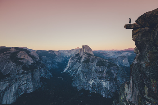 A fearless hiker is standing on an overhanging rock enjoying the view towards famous Half Dome at Glacier Point overlook in beautiful post sunset twilight, Yosemite National Park, California, USA