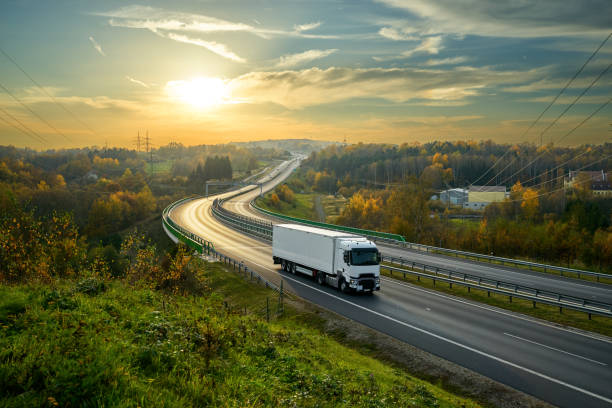 camion bianco che guida sull'autostrada che si snoda attraverso il paesaggio boscoso nei colori autunnali al tramonto - truck horizontal shipping road foto e immagini stock