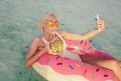 Woman floating in inflatable doughnut in ocean. She is enjoying and relaxing with cocktail. In right hand she is holding orange cocktail with straw in it. This sexy woman is wearing two pieces blue bikini and white sunglasses. Weather is perfect, there is some clouds but there is no worry about rain. She is relaxing and suntanning on warmth sunny summer day.