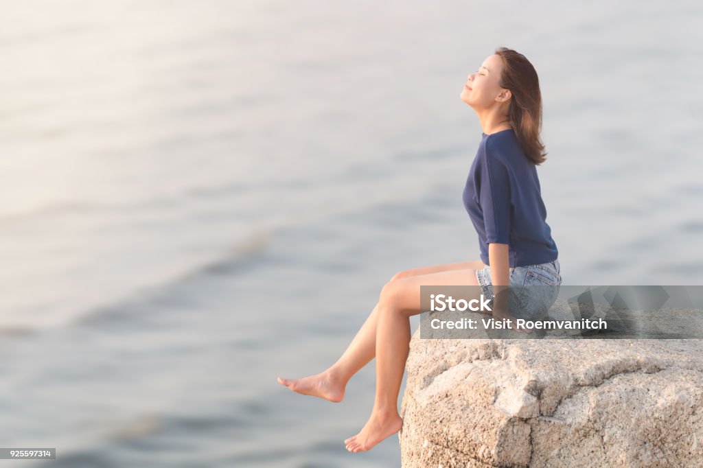 mujer con casual sentado en la roca acantilado relajando, sonreír y respirar profundo aire con borrosa mar ola - Foto de stock de Ejercicios de respiración libre de derechos