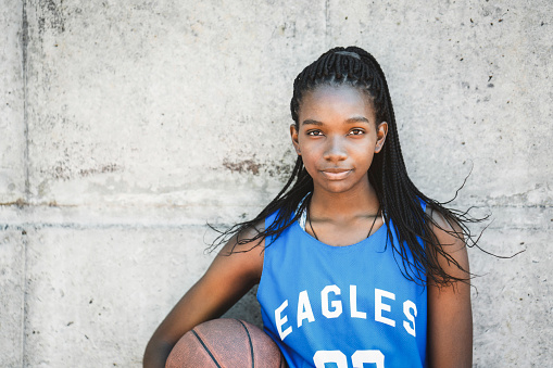 Female basketball player standing against wall. Confident teenage girl is holding ball. She is wearing blue jersey.