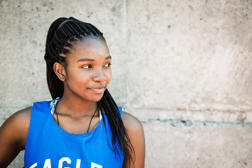 Confident female basketball player looking away. Teenage girl is standing against wall. She is wearing blue jersey.
