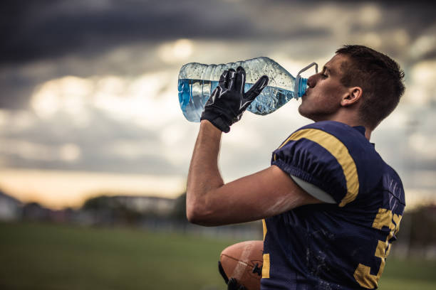 Thirsty American football player drinking fresh water on playing field. Profile view of young American football player drinking water on a break at playing field. groyne stock pictures, royalty-free photos & images