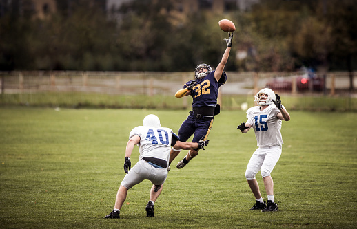 Determined American football player catching the ball between his rivals on a match at playing field.