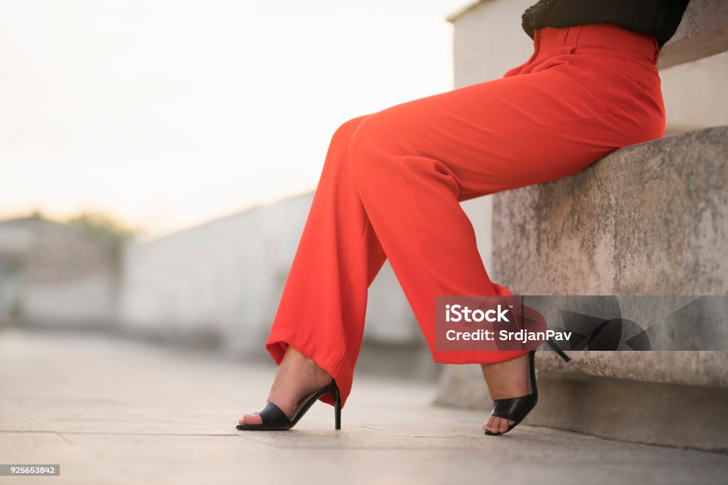 Summer In High Heels Fashion portrait of an unrecognizable young woman sitting at the balcony in red trousers and high heels; cut out. Pants Stock Photo