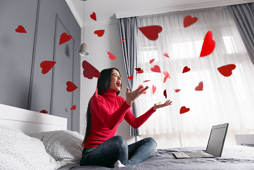Beautiful, happy, young woman throwing red heart-shapes in the air, sitting on the bed