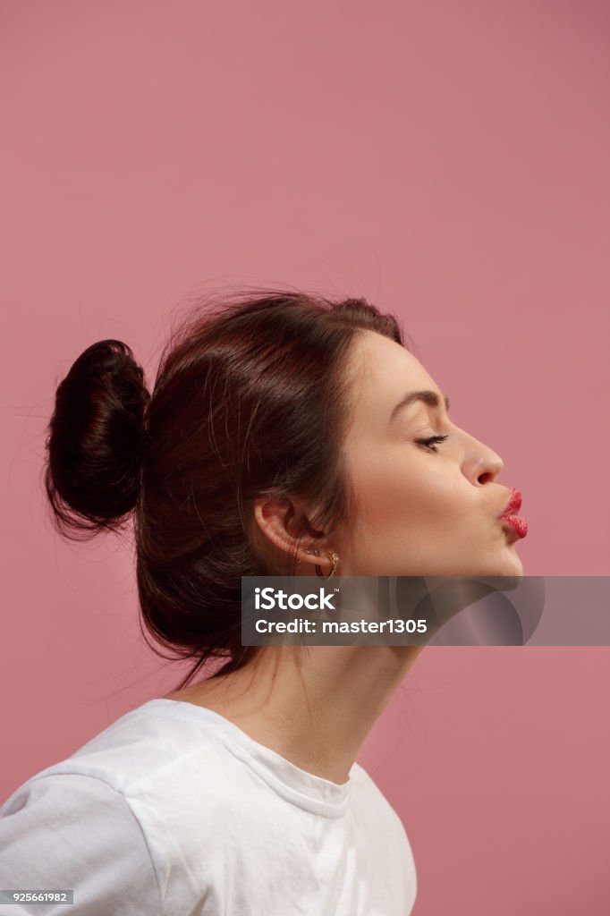 Portrait of attractive cute girl with bright makeup with kiss isolated over pink background I kiss you. Portrait of attractive woman with kiss on lips. Pink studio. Beautiful female portrait. Young happy emotional funny woman looking at camera. Human facial emotions concept. Profile Blowing Stock Photo