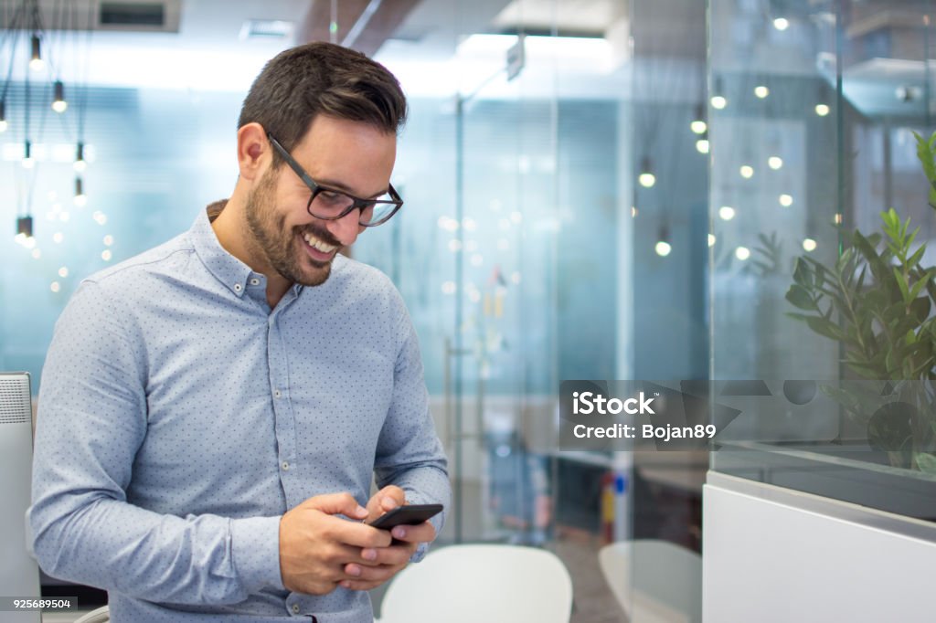 Smiling cheerful young man in smart casual wear holding smart phone and looking at it while standing in modern office. Mobile Phone Stock Photo