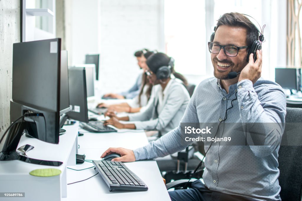 Operador de soporte cliente guapo sonriente con auriculares trabajando en call center. - Foto de stock de Agente de servicio al cliente libre de derechos