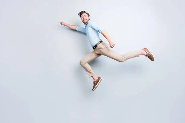 Photo of Happy, attractive, handsome, young man with bristle jumping in air showing superman pose looking at camera with beaming smile over grey background