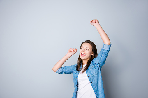 Portrait of happy cute young woman with toothy smile in casual oufit, raised hands, dancing because her dream comes true, standing over grey background