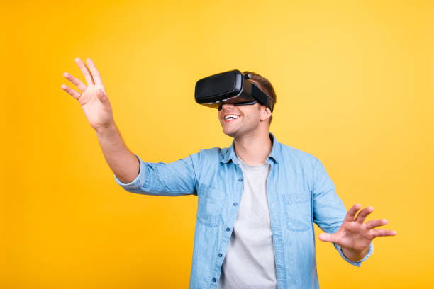 istock Cheerful, glad guy in jeans shirt wearing virtual reality goggles, getting experience using VR-headset, gesticulating, touching something with arms while standing over yellow background 925694128