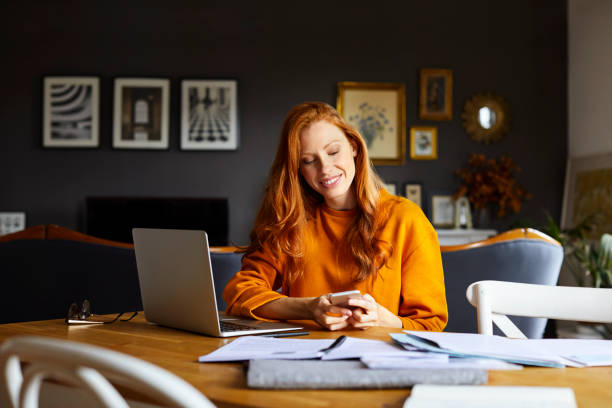 Smiling businesswoman using mobile phone at home Smiling businesswoman using mobile phone at home. Young female is with laptop and documents. She is sitting at table. redhead stock pictures, royalty-free photos & images