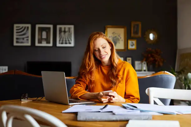 Smiling businesswoman using mobile phone at home. Young female is with laptop and documents. She is sitting at table.