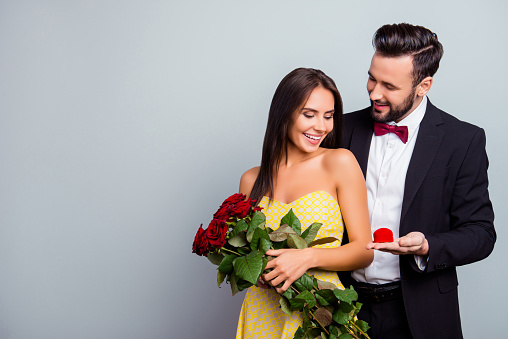 Portrait with copy space of attractive family, couple in formal wear, sexy wife in dress holding bouquet of red roses, husband in tuxedo embracing her from back side and presenting a case with ring