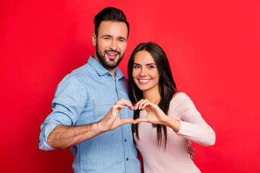 Concept photo of love story of cheerful, attractive, lovely, cute, sweet couple in casual outfit making heart with fingers over red background, ideal wife and husband on 14 february