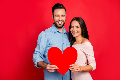 He vs she celebrating valentine day. Portrait of caucasian, beautiful, lovely, cute, cheerful, positive couple holding red paper heart together standing over red background
