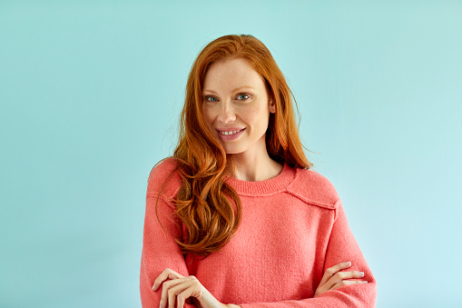 Portrait of smiling woman with long hair. Beautiful young female is standing with arms crossed against blue background. She is wearing sweater.