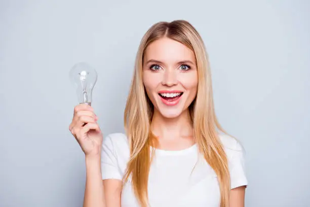 Photo of Wow! It is so easy! Close up portrait of cute lovely clever confident excited beautiful woman with blonde hair with open mouth, she is holding a light bulb, isolated on grey background