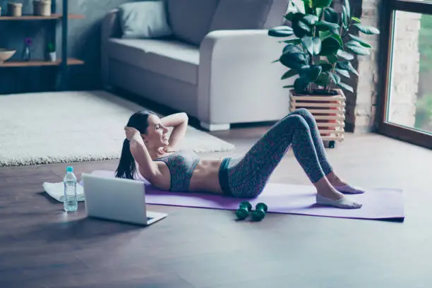 Purposeful strong athletic sportive beautiful attractive sportswoman lying on the floor and doing crunches stretching abs, she is imitating video lessons