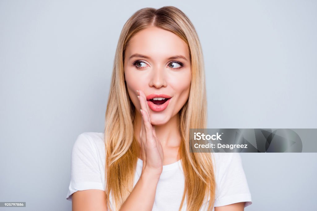 ¡No decir a nadie! Hablador misteriosa mujer muy hermosa con cabello rubio, vestido con la camiseta blanca es decir secreto caliente noticias de frenado y mirando a un lado, aislado en fondo gris - Foto de stock de Susurrar libre de derechos